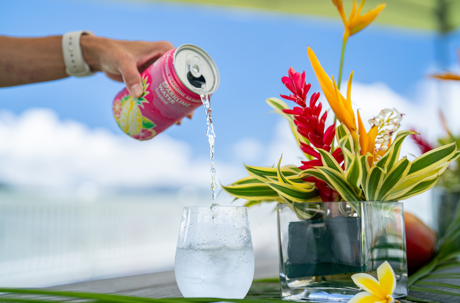 a promotional image that shows a hand pouring a tropical sparkling water into a glass. a nice tropical flower arrangement is on the side.